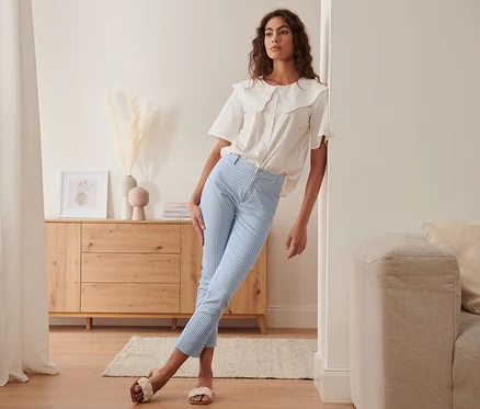 Femme appuyée contre un mur portant un pantalon stretch à rayures et un chemisier blanc.
