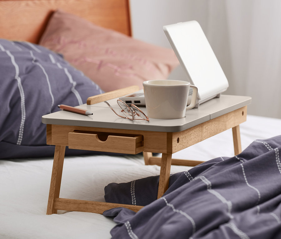 Sur un lit, une table de lit avec un ordinateur portable, une tasse et des lunettes.
