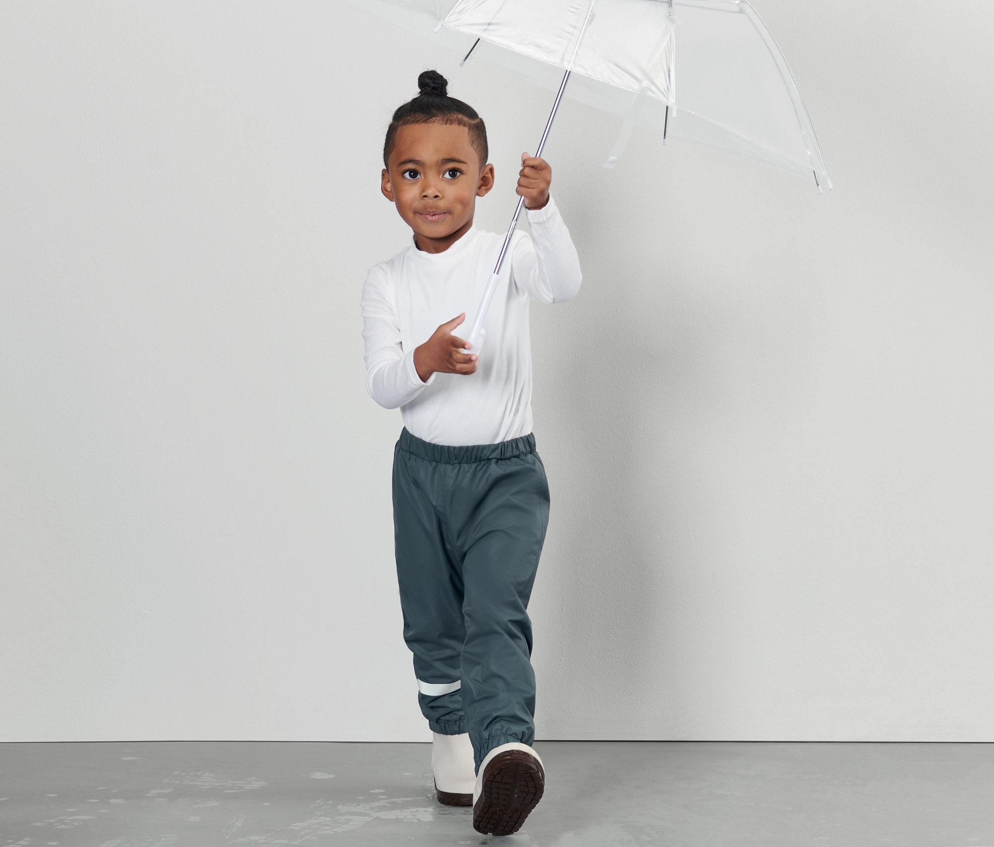 Un garçon tient un parapluie transparent et porte un pantalon imperméable pour tout-petit.