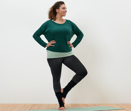 Une femme debout sur un tapis de yoga porte un t-shirt de sport 2 en 1 vert, un legging de sport et des collants noirs.
