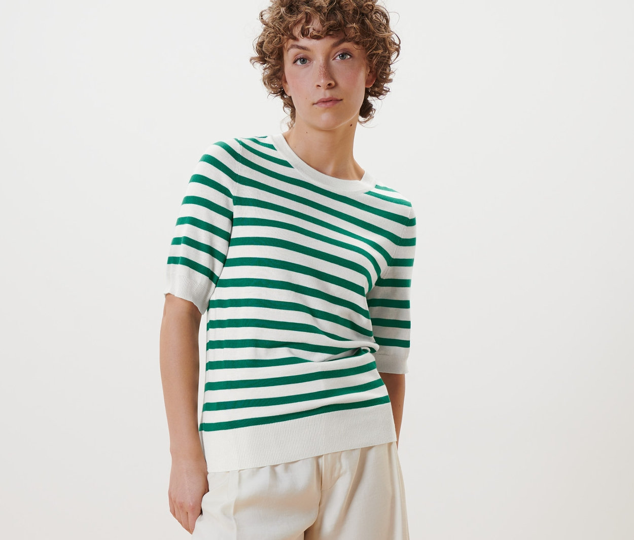 Femme posant dans un pull en maille fine rayé, un pantalon blanc et des chaussures blanches.