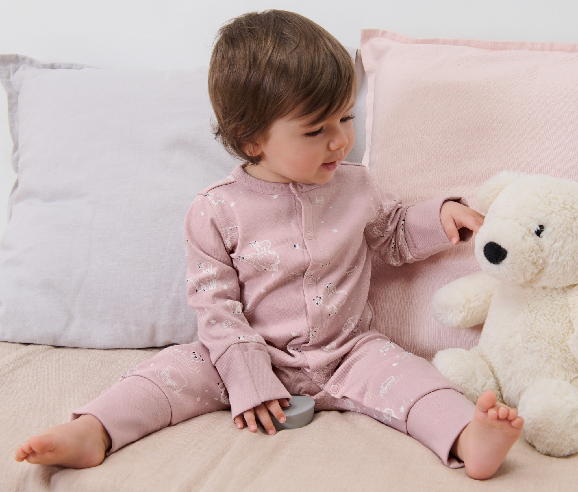 Un enfant est assis sur un lit et joue avec un ours en peluche. Il porte un ensemble de 3 pyjamas roses pour bébé.