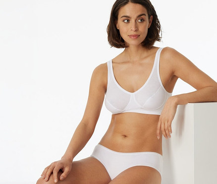 Une femme aux cheveux courts et foncés porte une brassière SCHIESSER blanche à bonnets et une culotte blanche, assise sur un bloc blanc et appuyée sur un autre.