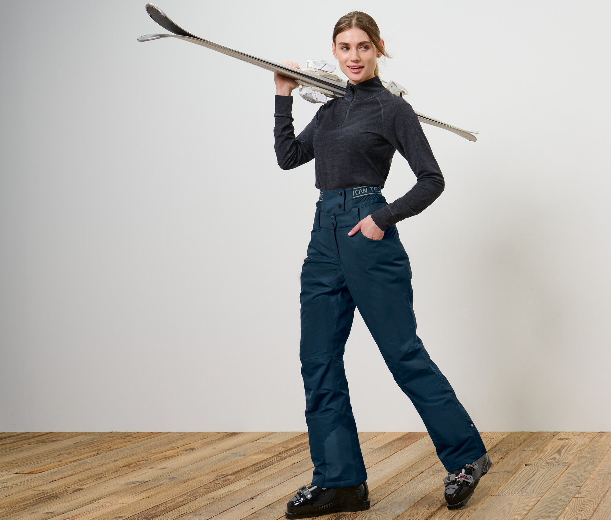 Femme posant en pantalon de ski et de snowboard bleu marine, tenant des skis sur l'épaule.