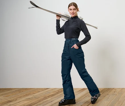 Femme posant en pantalon de ski et de snowboard bleu marine, tenant des skis sur l'épaule.