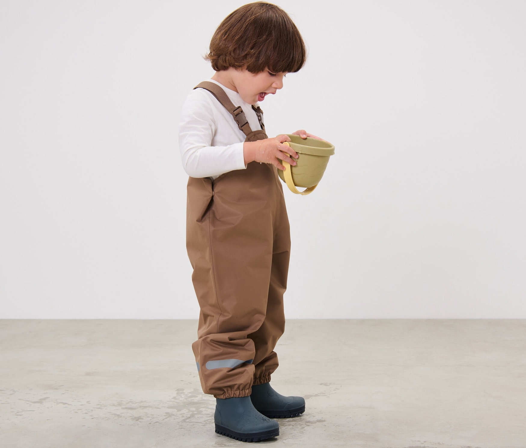 Un enfant portant un pantalon de pluie marron et des bottes en caoutchouc bleues tient un seau vert.