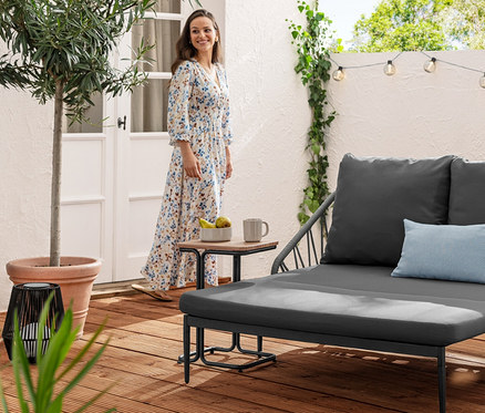 Une femme en robe se tient près d'une table C en aluminium à côté d'un canapé lounge extensible. À l'arrière-plan, on peut voir une guirlande lumineuse solaire avec des boules rainurées.