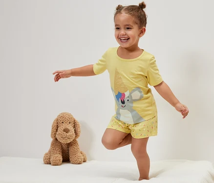 Une fillette souriante se tient debout sur un lit à côté d'un chien en peluche et porte un pyjama court jaune pour enfant, koala.