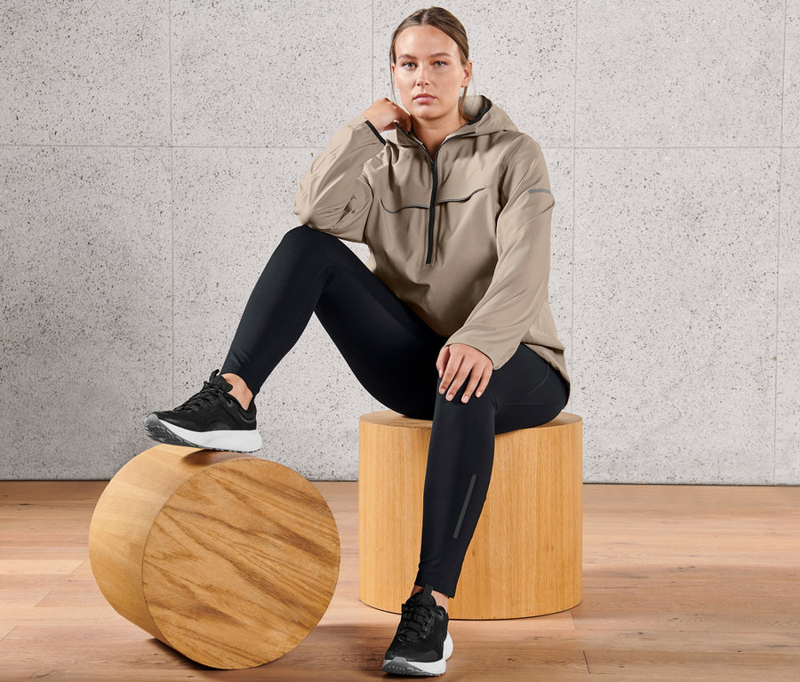 Femme assise sur un tabouret en bois portant une veste de sport imperméable taupe et un legging de sport haute performance.