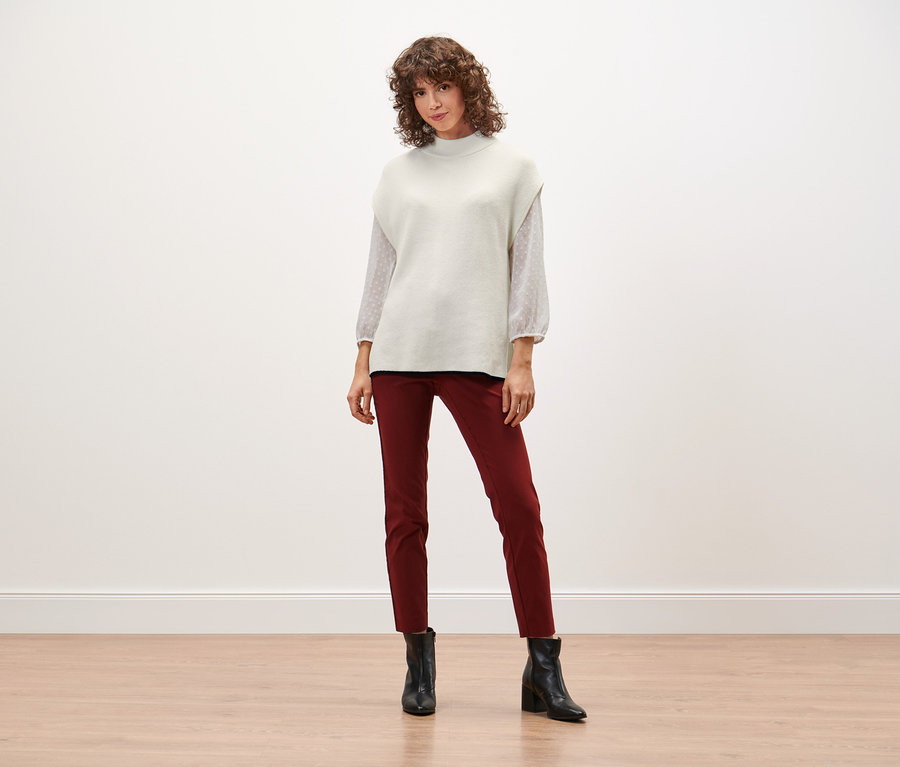 Une femme debout portant un pull sans manches en maille, un pantalon rouge et des bottes noires.