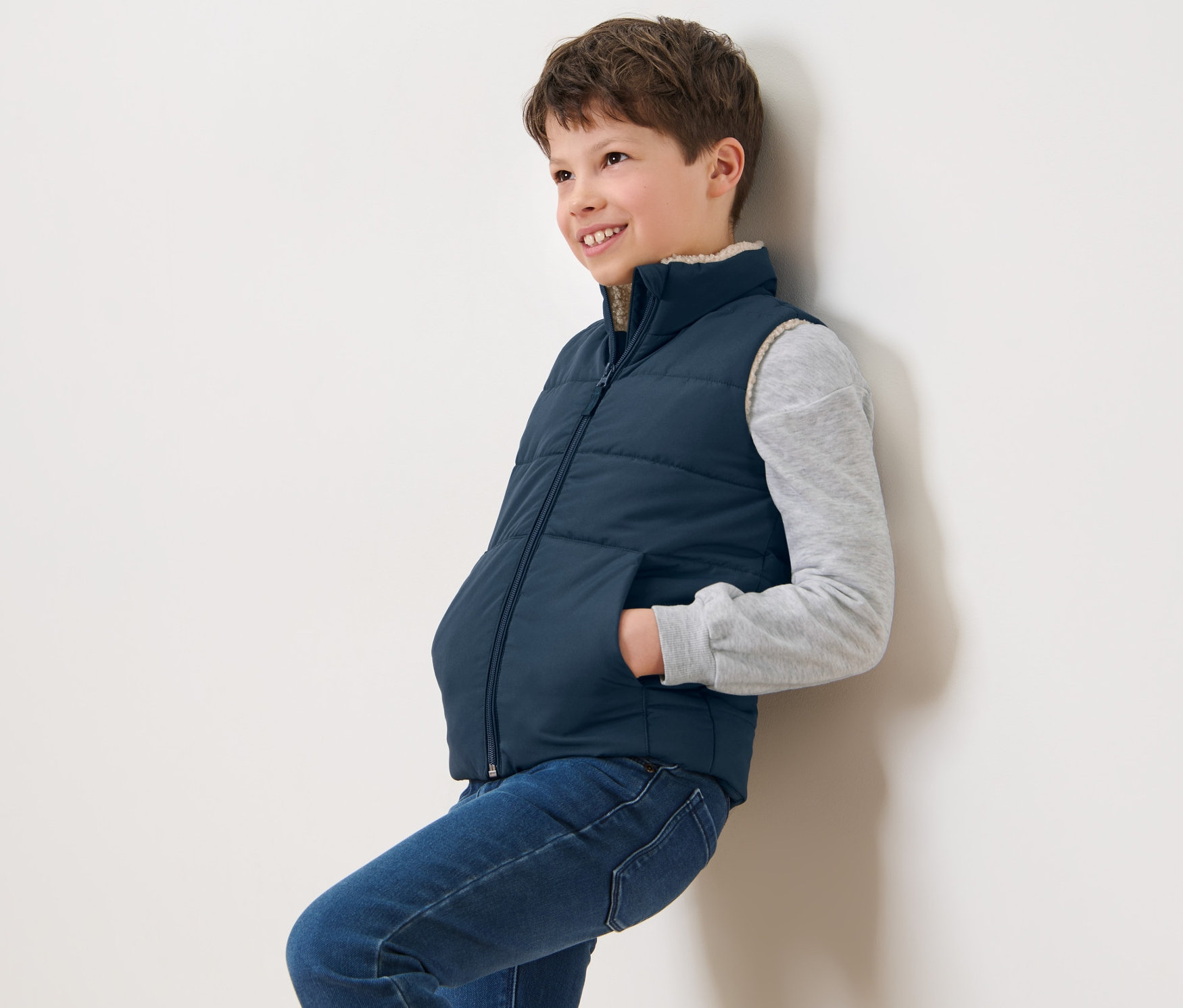 Un garçon appuyé contre un mur porte un gilet sans manches matelassé en teddy bleu marine pour enfant.