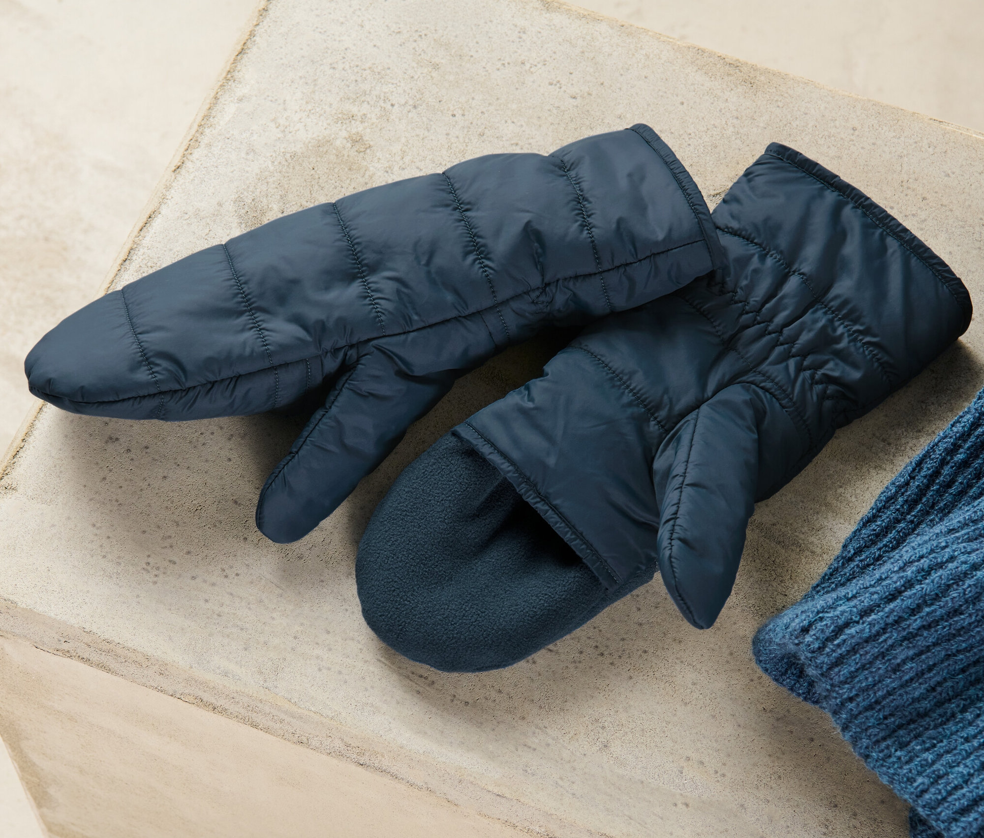 Deux mitaines-moufles matelassées bleu marine sont posées sur une surface grise à côté d'un bonnet bleu en tricot.