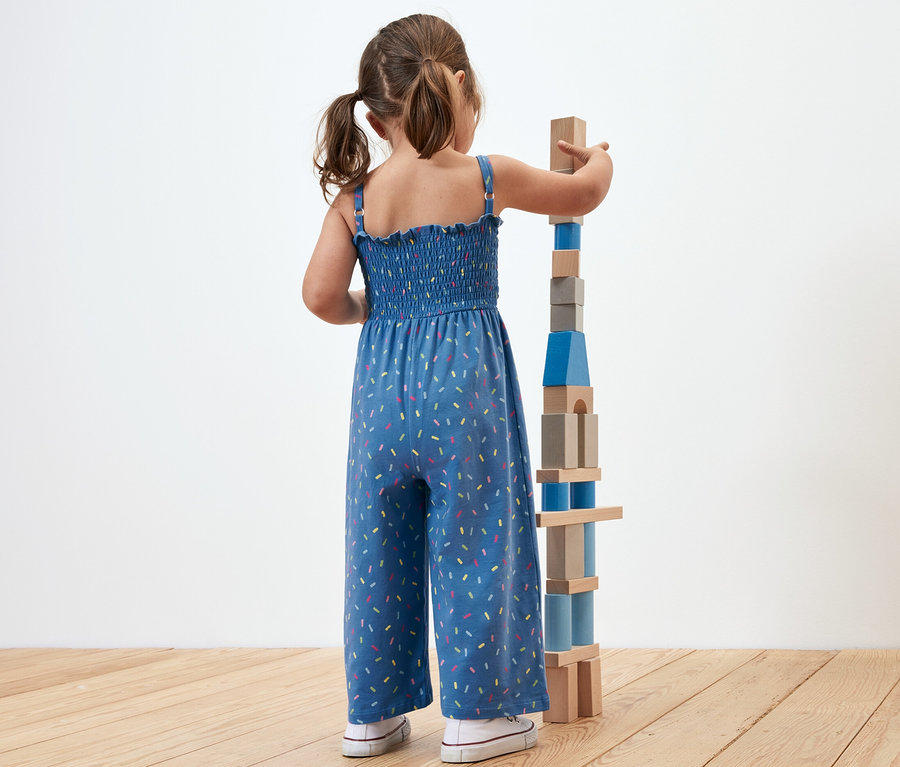 Fillette construisant une tour de blocs de bois. Elle porte une combinaison de longueur 7/8.