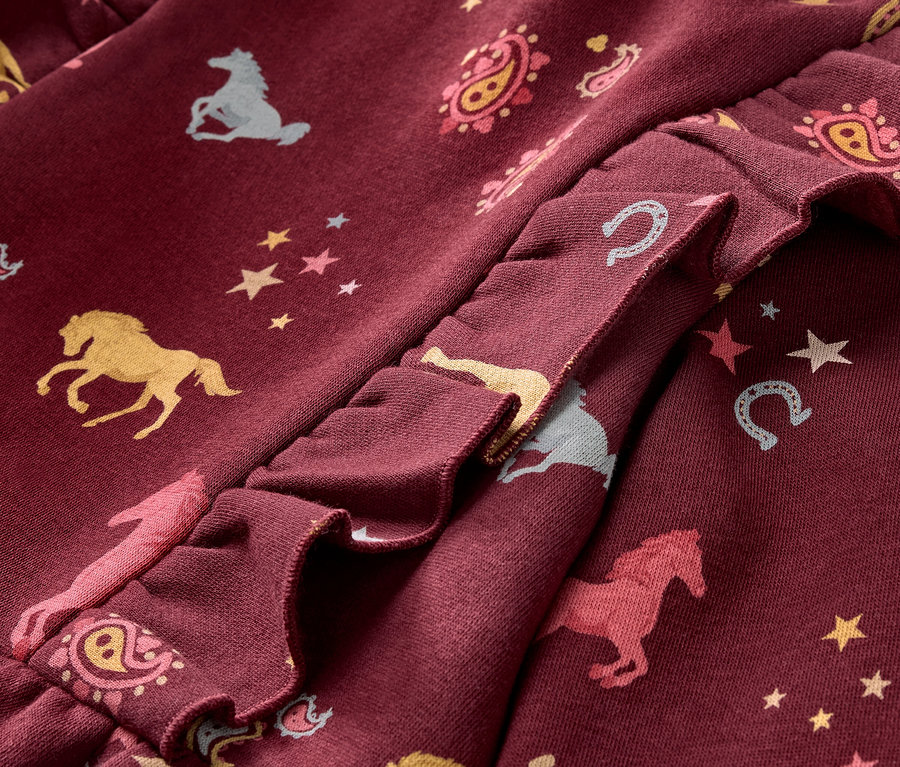 Gros plan sur une robe en sweat bordeaux pour enfant, avec des volants et des motifs de chevaux, d'étoiles et de fers à cheval.