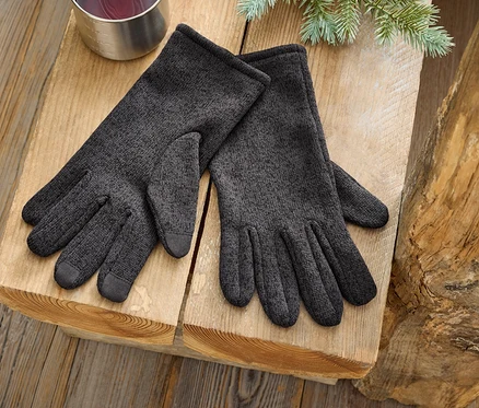Une paire de gants gris est posée sur une surface en bois à côté d'une tasse argentée et d'une branche de conifère.