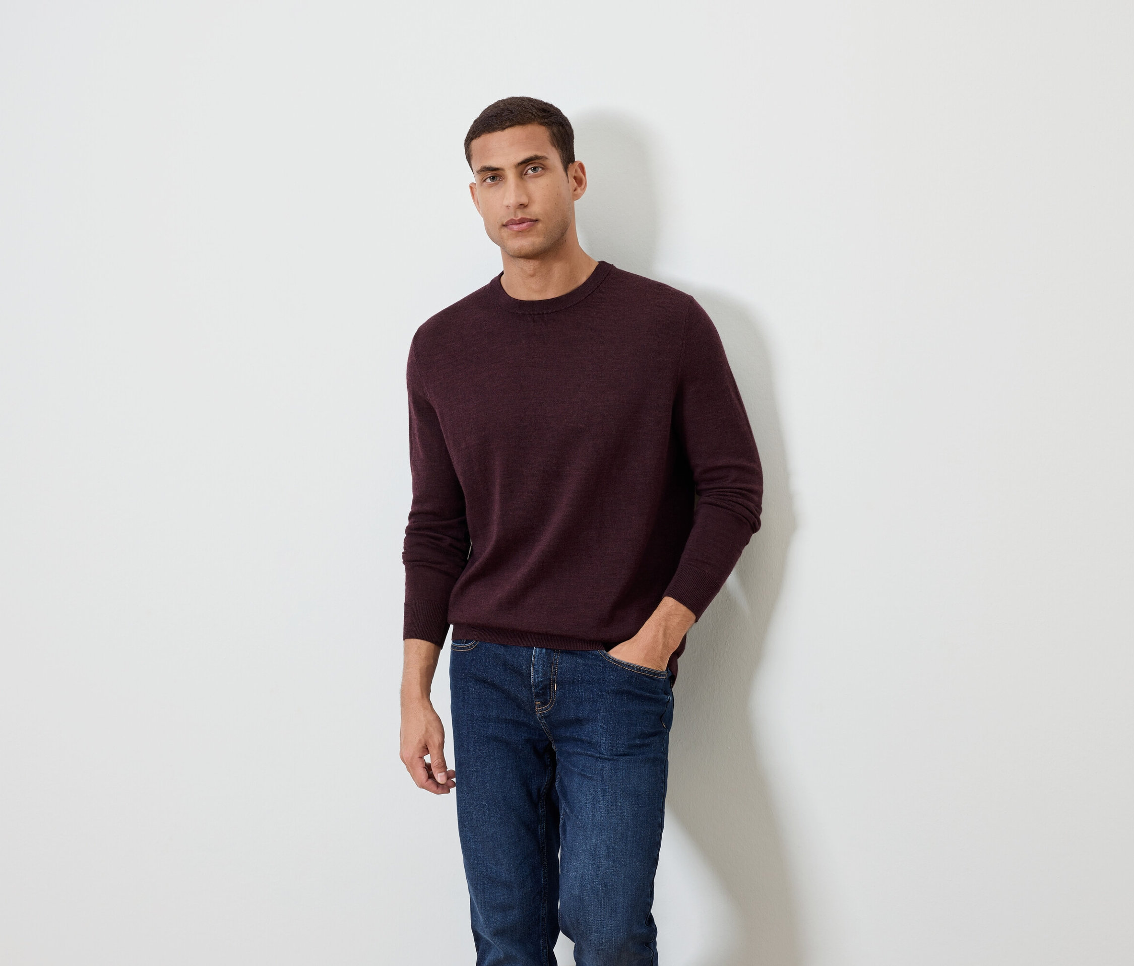 Un homme pose sur un fond blanc, portant un pull bordeaux et un jean.