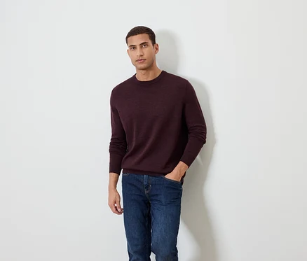 Un homme pose sur un fond blanc, portant un pull bordeaux et un jean.