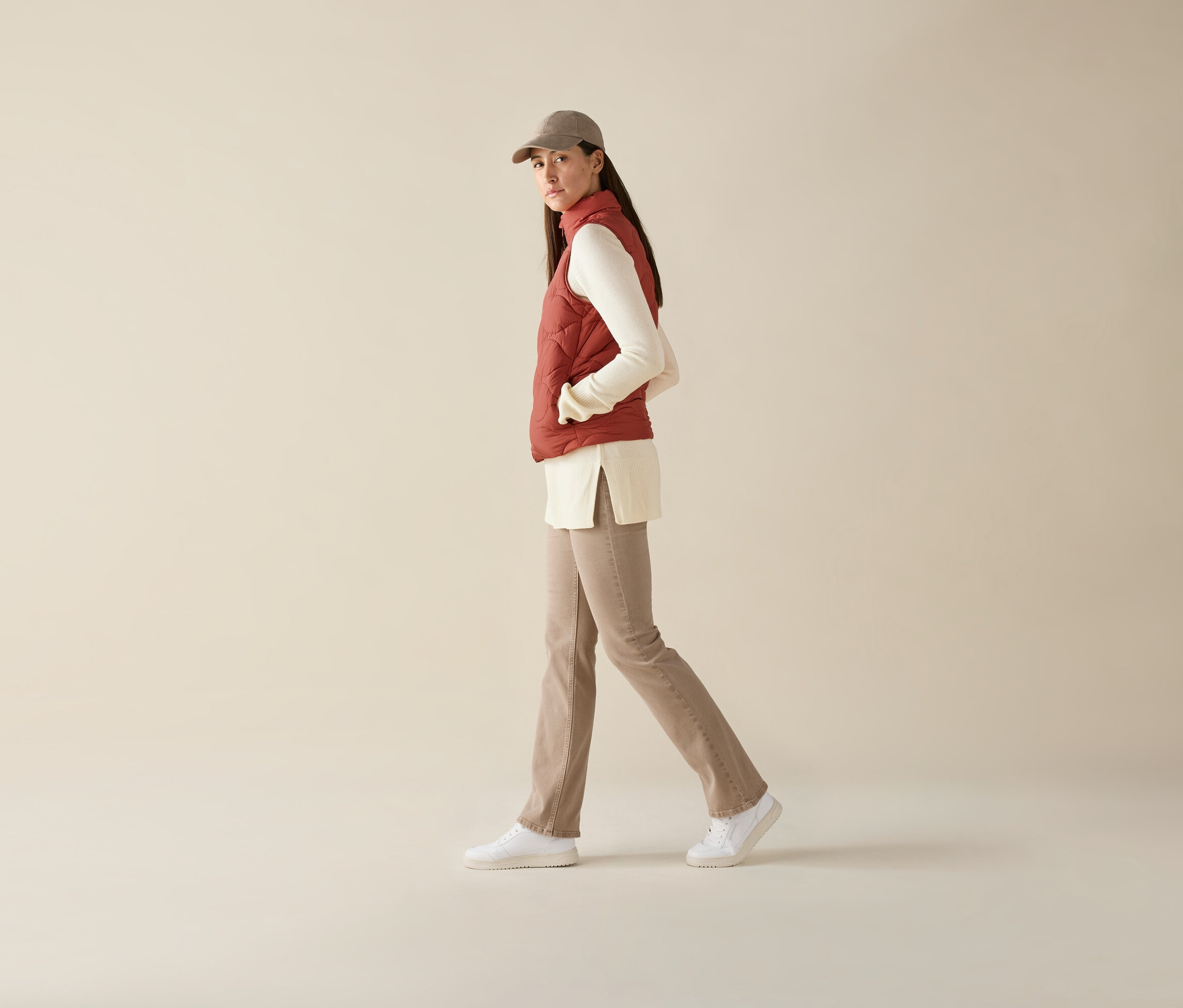 Femme marchant portant un gilet matelassé sans manches, un haut à manches longues, un pantalon et une casquette de baseball.