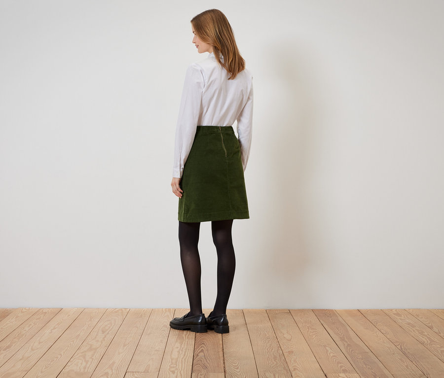Une femme se tient dos à la caméra et porte une jupe en velours côtelé verte, une chemise blanche, des collants noirs et des chaussures noires.