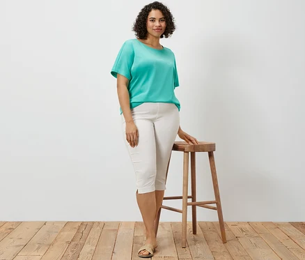 Une femme pose, appuyée sur un tabouret en bois, portant un pantalon 3/4 stretch blanc et un chemisier turquoise à encolure dégagée.