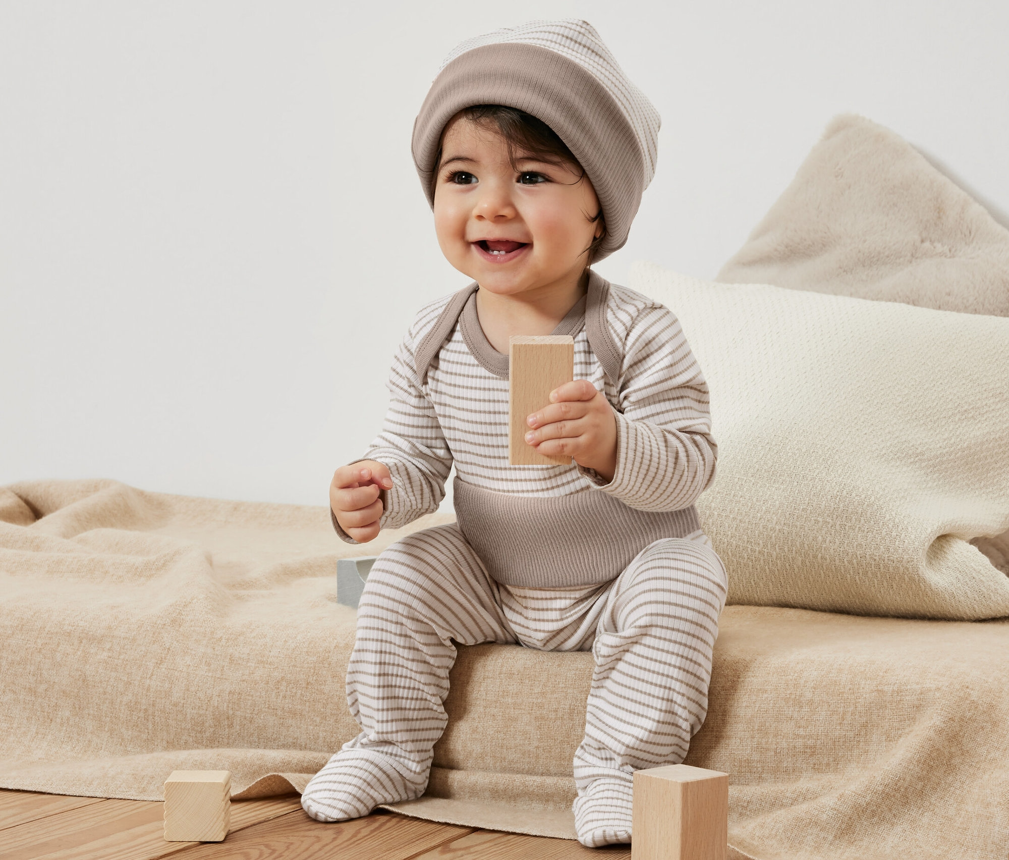 Un bébé souriant est assis sur une couverture et joue avec des blocs de bois. Il porte un ensemble pour bébé rayé, 3 pièces.