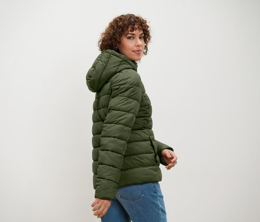 Femme aux cheveux bouclés regardant par-dessus son épaule posant dans une veste matelassée vert olive à capuche et une veste en jean.