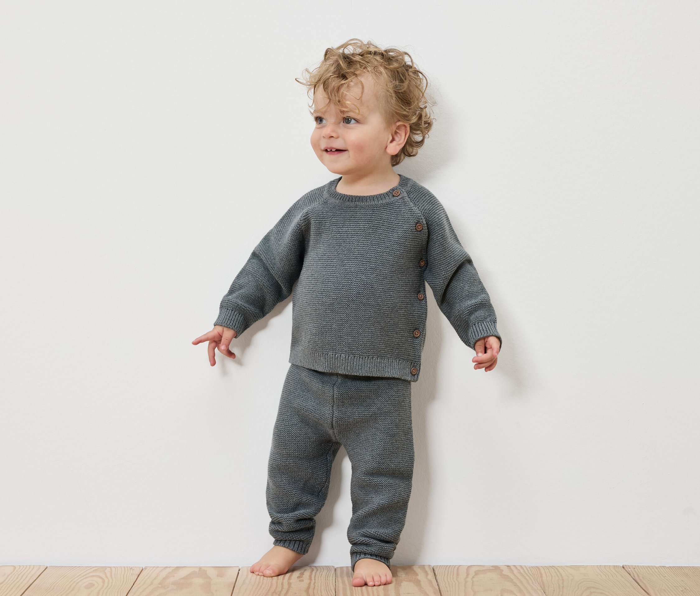 Un jeune enfant debout devant un mur blanc porte un ensemble en maille pour bébé.
