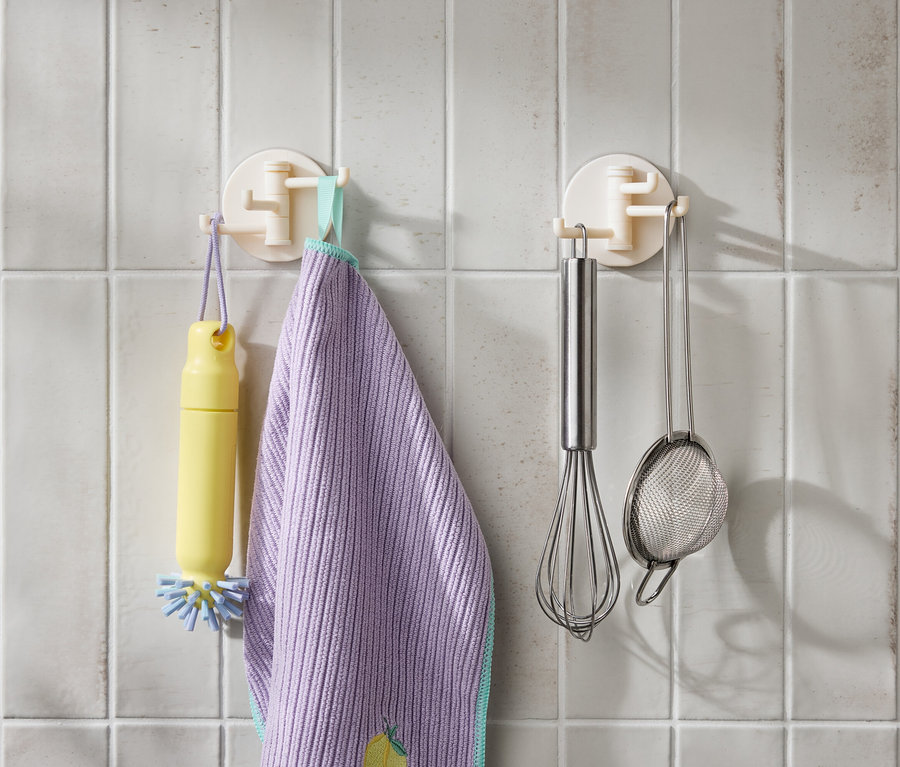 Sur des carreaux blancs sont accrochés une brosse jaune, un torchon violet, un fouet en inox et une passoire.