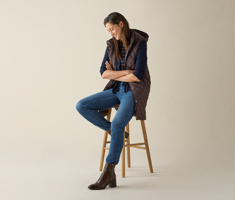 Une femme est assise sur un tabouret, les bras croisés, et porte un long gilet matelassé à capuche marron foncé.