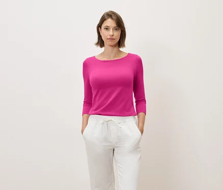Une femme aux cheveux bruns pose dans un haut rose à manches trois-quarts et un pantalon blanc.