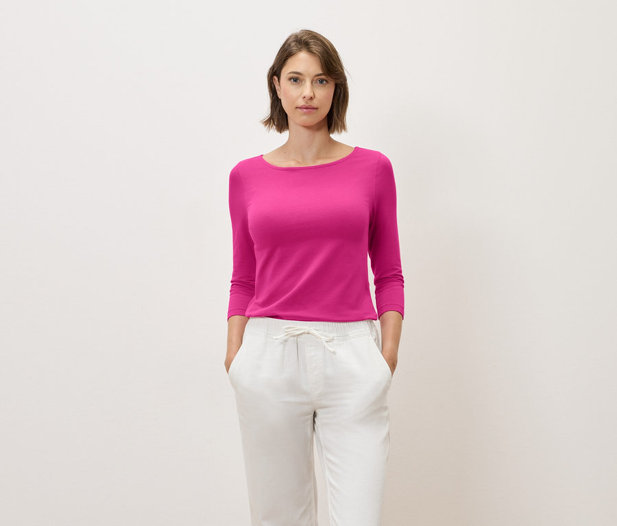 Une femme aux cheveux bruns pose dans un haut rose à manches trois-quarts et un pantalon blanc.