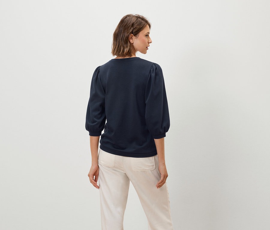 Une femme porte un haut bleu marine à manches trois-quarts et un pantalon beige.