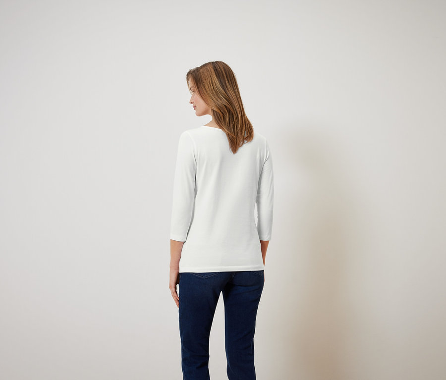 Une femme aux cheveux bruns, vue de dos, porte un t-shirt blanc à manches trois-quarts et un jean bleu.