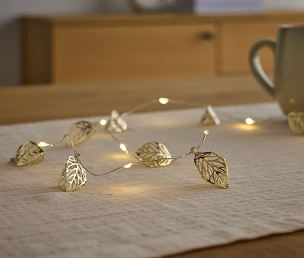 Une guirlande lumineuse dorée en forme de feuilles repose sur un chemin de table à côté d'une tasse.
