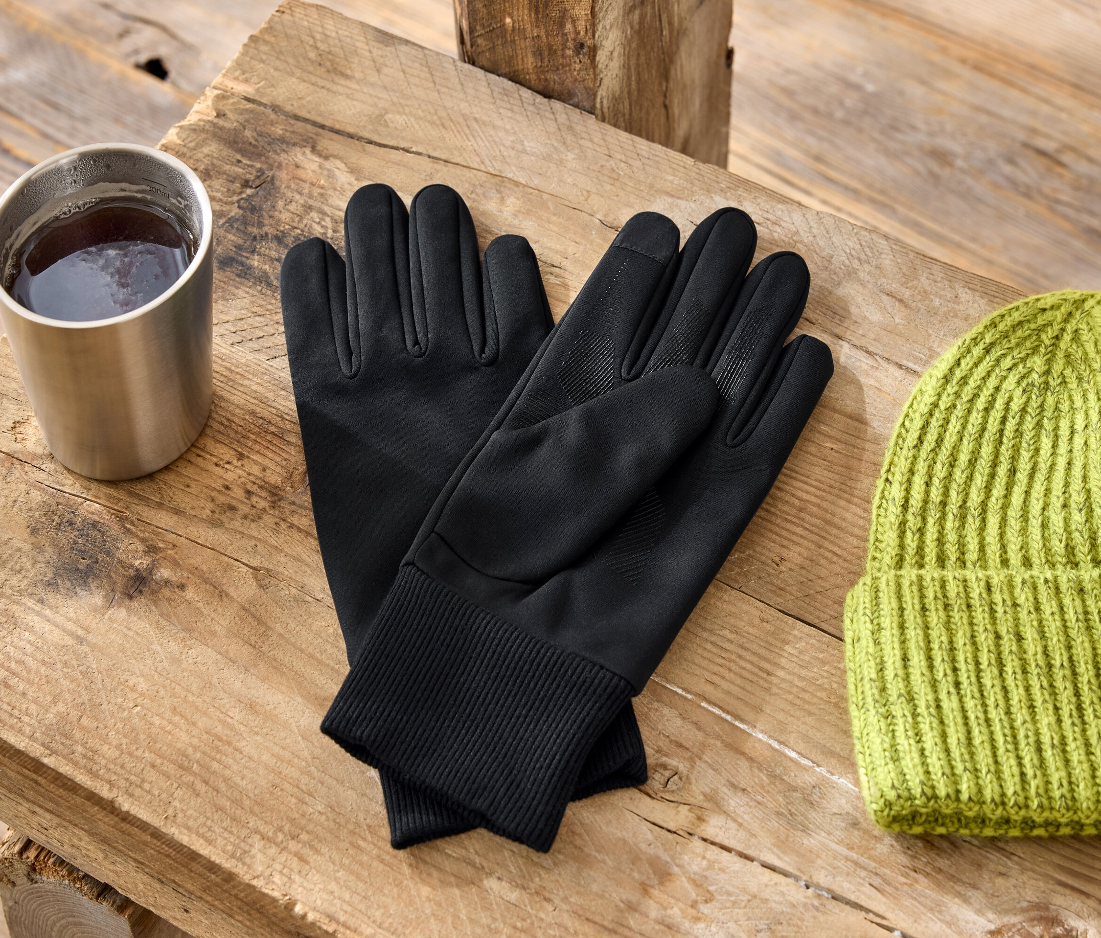 Sur une table en bois se trouvent une tasse en métal, des gants noirs et un bonnet tricoté vert.