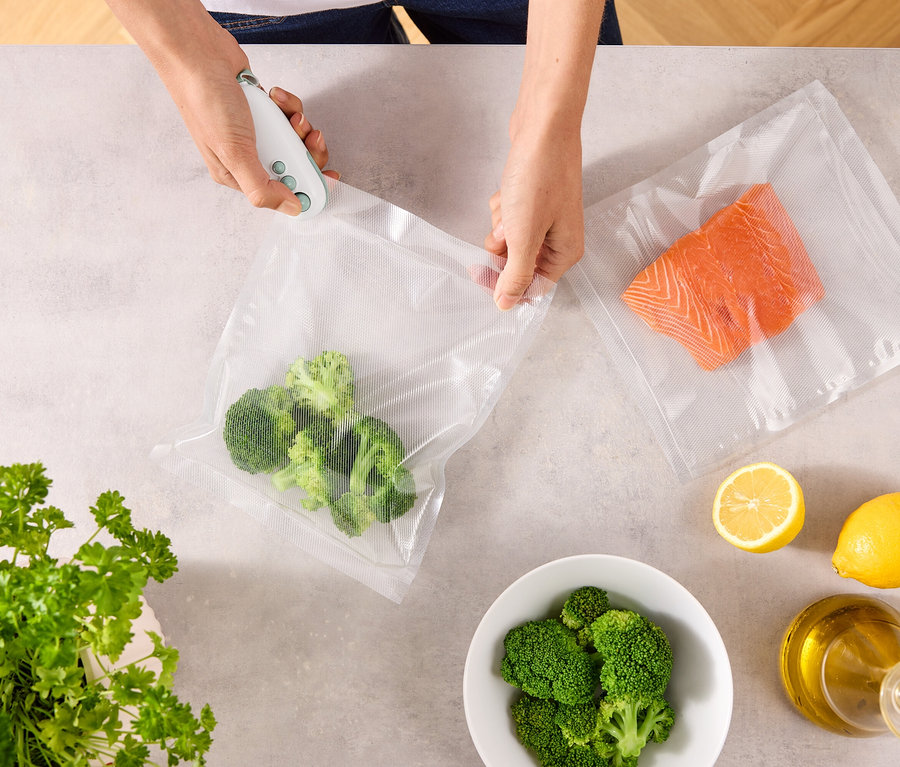 Une personne scelle un sac de brocolis avec un appareil de mise sous vide. Sur la table, il y a un autre sac avec du saumon, des citrons, un bol de brocolis, du persil et une bouteille d'huile.