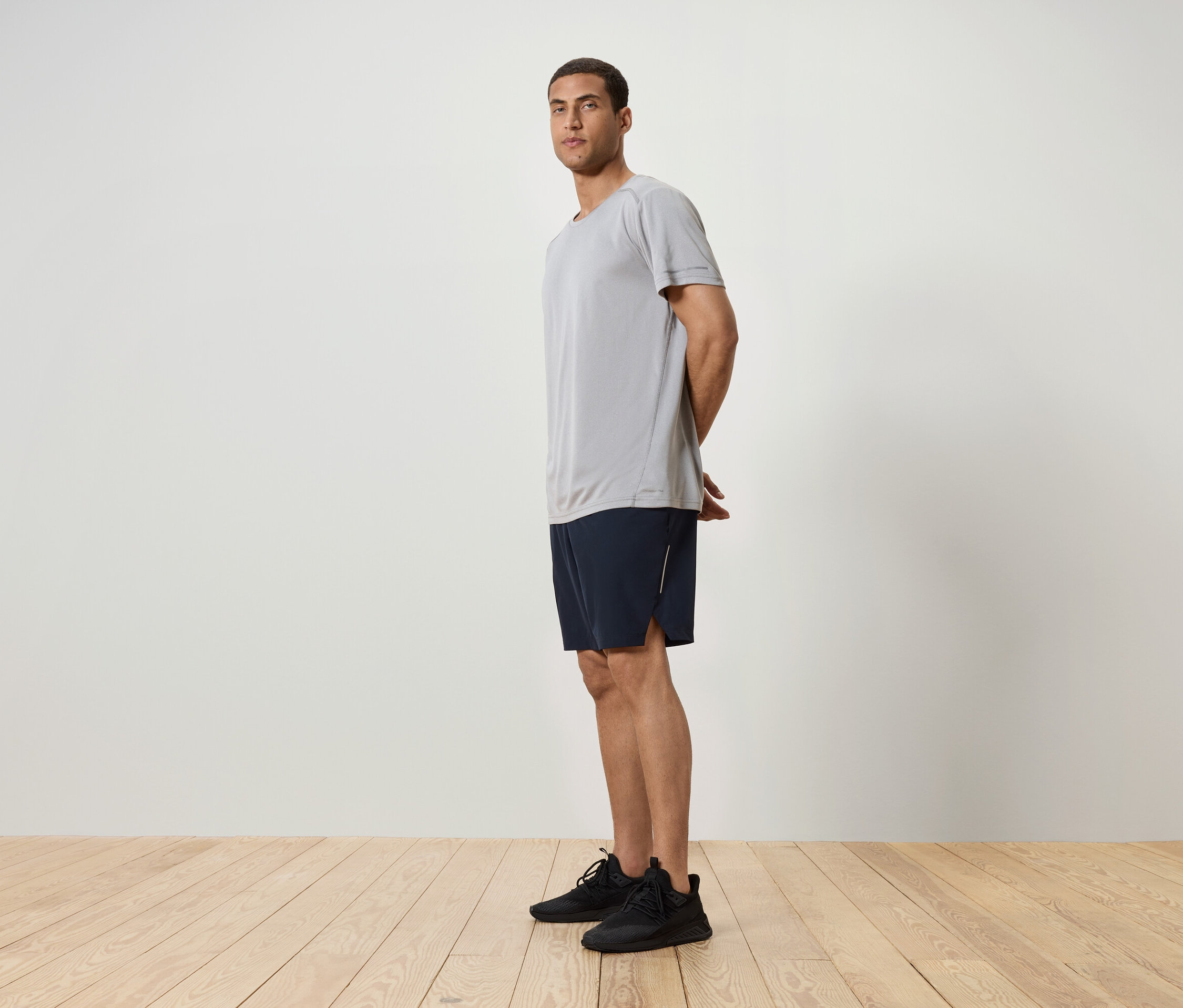 Un homme se tient de profil, portant un t-shirt gris, un short bleu marine et des baskets noires.