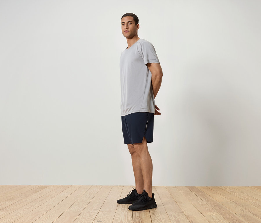 Un homme se tient de profil, portant un t-shirt gris, un short bleu marine et des baskets noires.