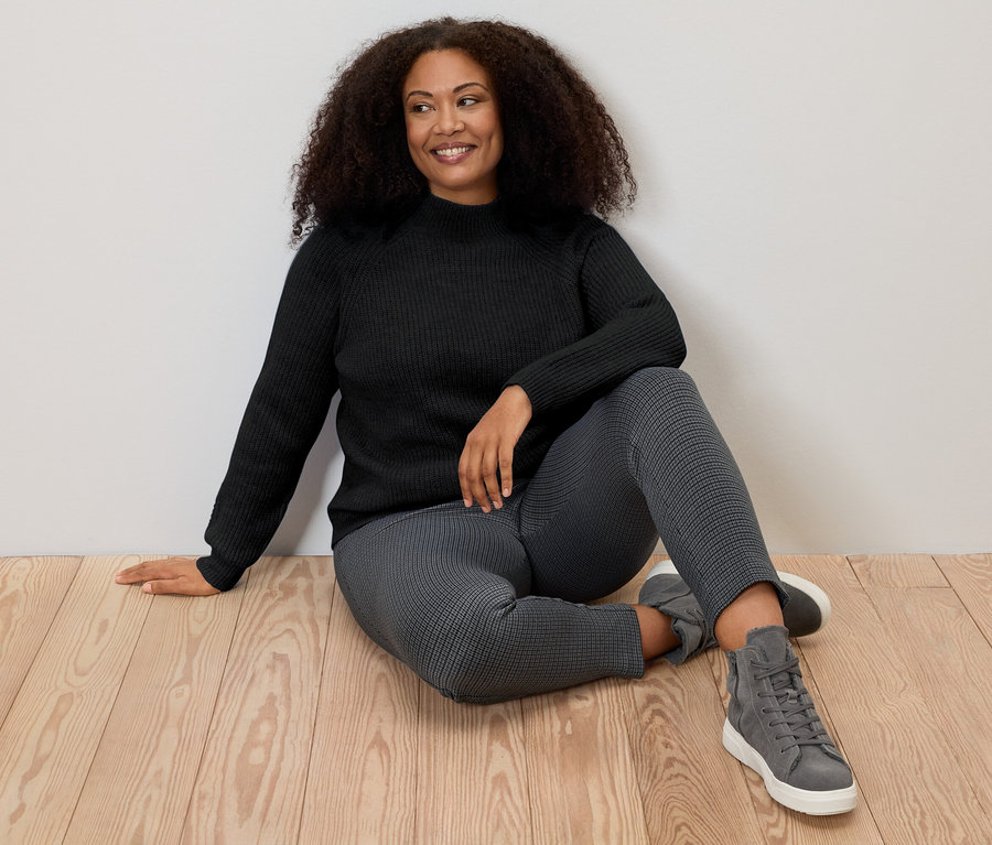 Femme assise par terre portant un pull noir à col droit en grosse maille.