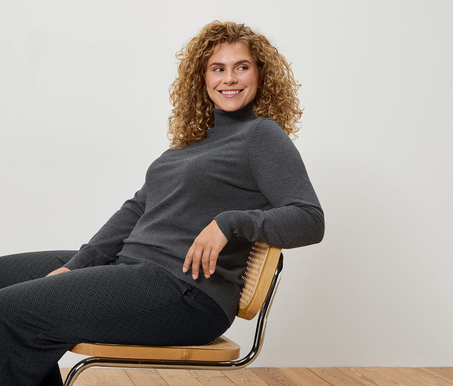 Une femme aux cheveux bouclés est assise sur une chaise et porte un col roulé gris et un pantalon à motifs.