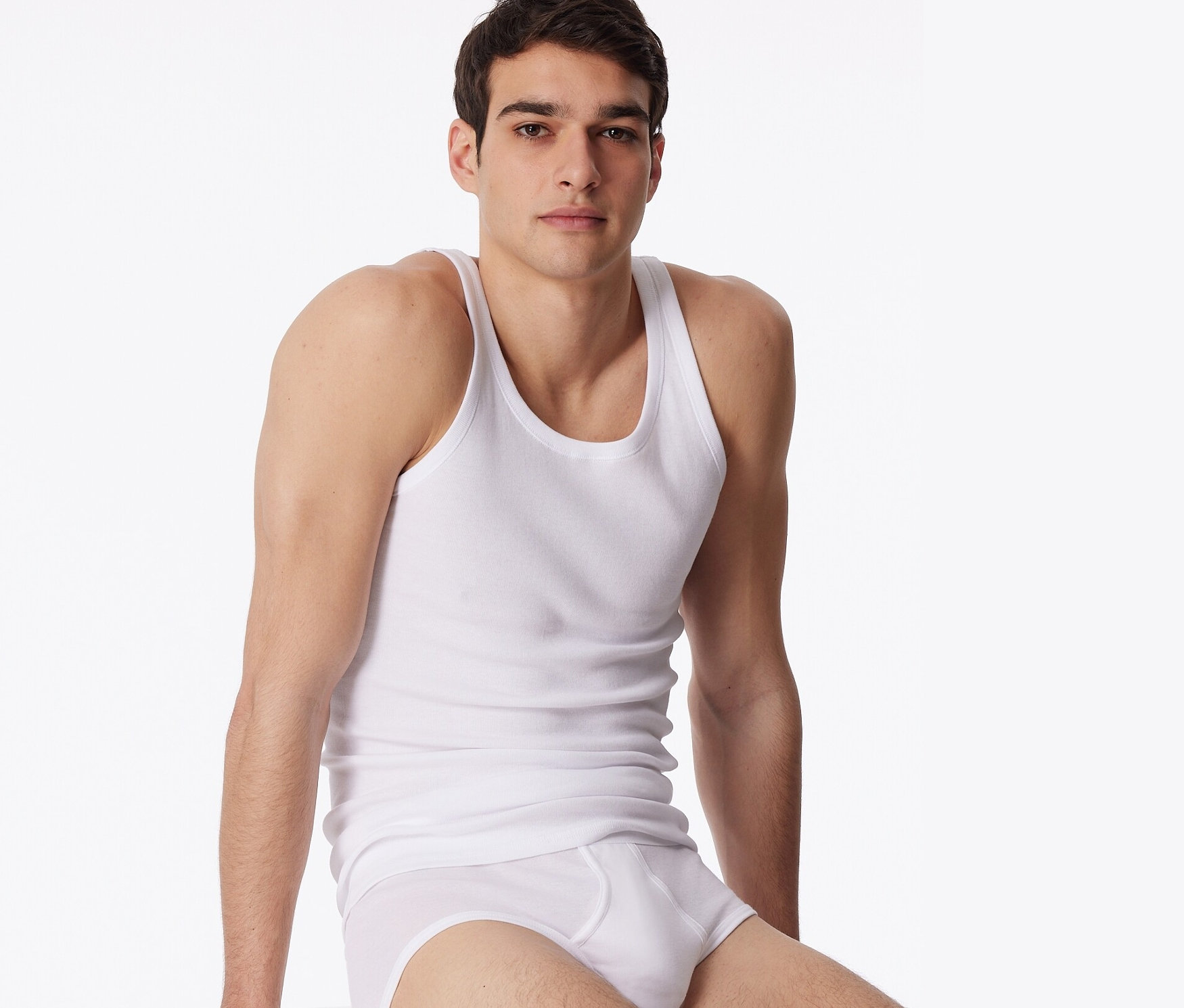 Un homme est assis sur un cube blanc et porte des sous-vêtements blancs et un lot de 2 maillots de corps blancs SCHIESSER en maille côtelée fine.