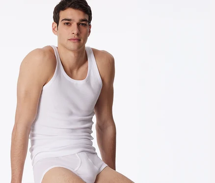 Un homme porte un maillot de corps SCHIESSER blanc en maille côtelée fine et un slip blanc, assis sur un cube blanc.