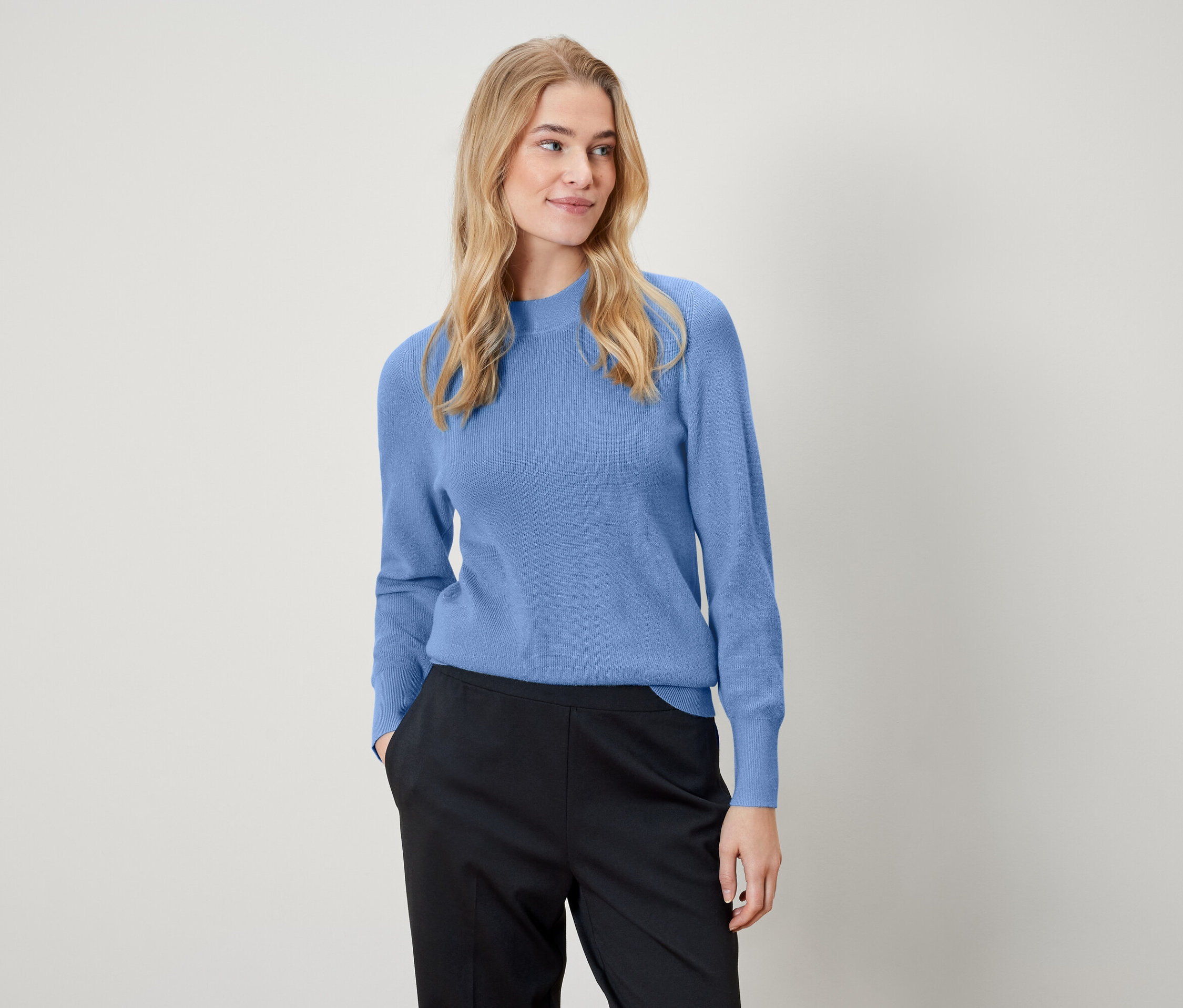 Une femme blonde porte un pull bleu clair et un pantalon noir.
