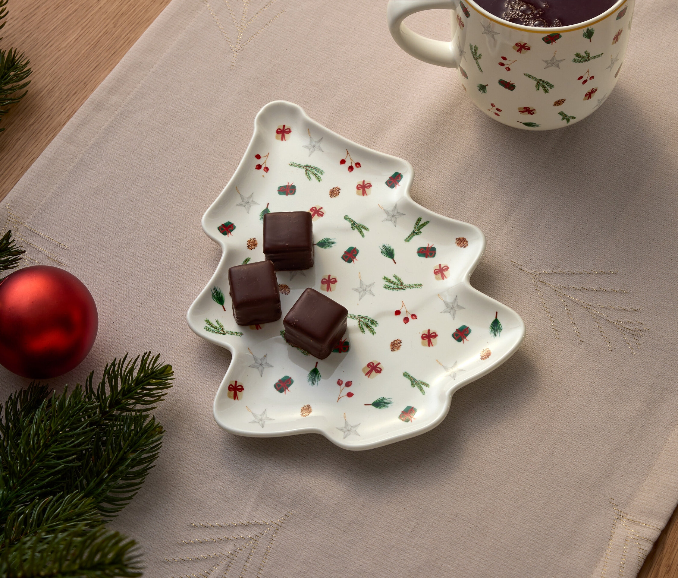 Trois chocolats sur une assiette en forme de sapin de Noël, à côté d'une tasse et d'une décoration de Noël.