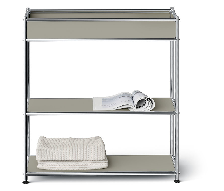 Étagère à trois niveaux avec cadre argenté et étagères grises. Un magazine ouvert est posé sur l'étagère du milieu, une couverture blanche pliée est posée sur l'étagère du bas.