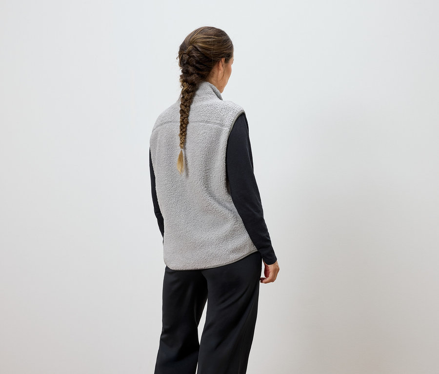 Femme vue de dos avec une tresse, portant un gilet gris et un pantalon noir.