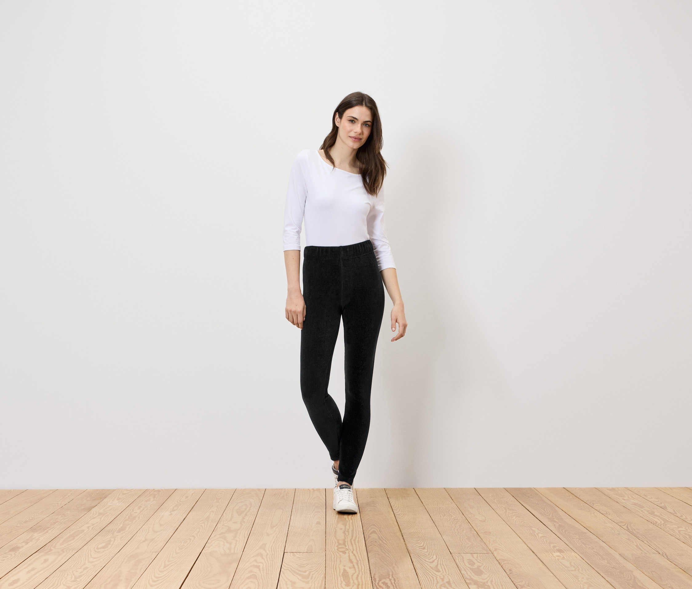 Une femme se tient sur un plancher en bois, portant un chemisier blanc et un pantalon noir.