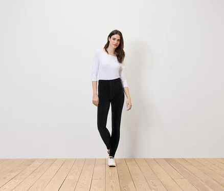 Une femme se tient sur un plancher en bois, portant un chemisier blanc et un pantalon noir.
