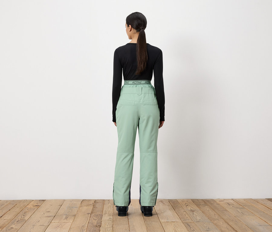 Une femme, de dos, porte un t-shirt noir à manches longues et un pantalon vert clair.