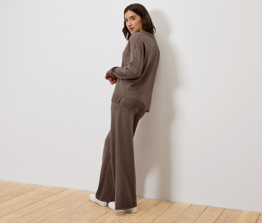 Une femme appuyée contre un mur porte un haut et un pantalon en tricot marron.